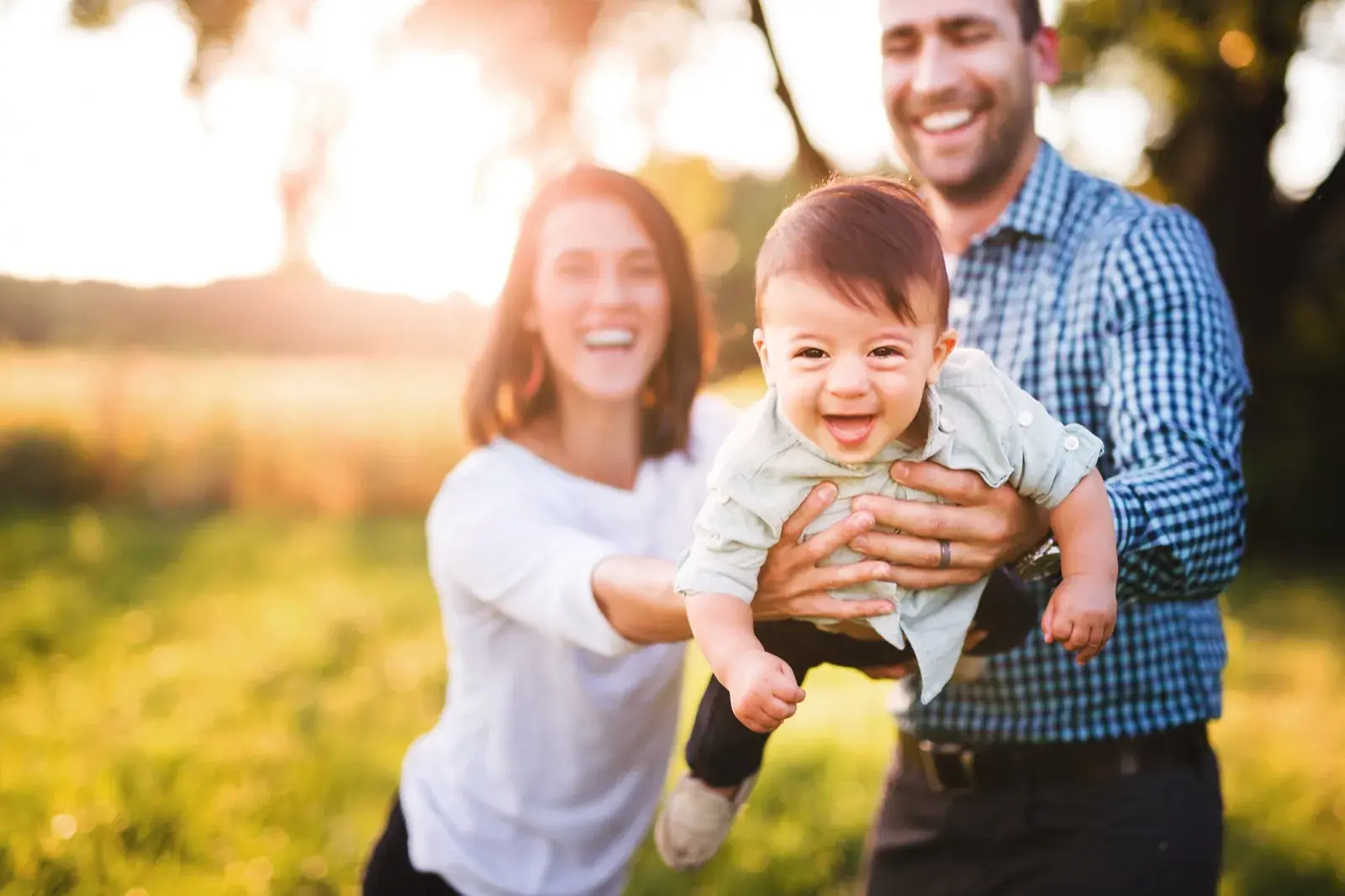 Parents laughing as they lift their baby into the air outdoors